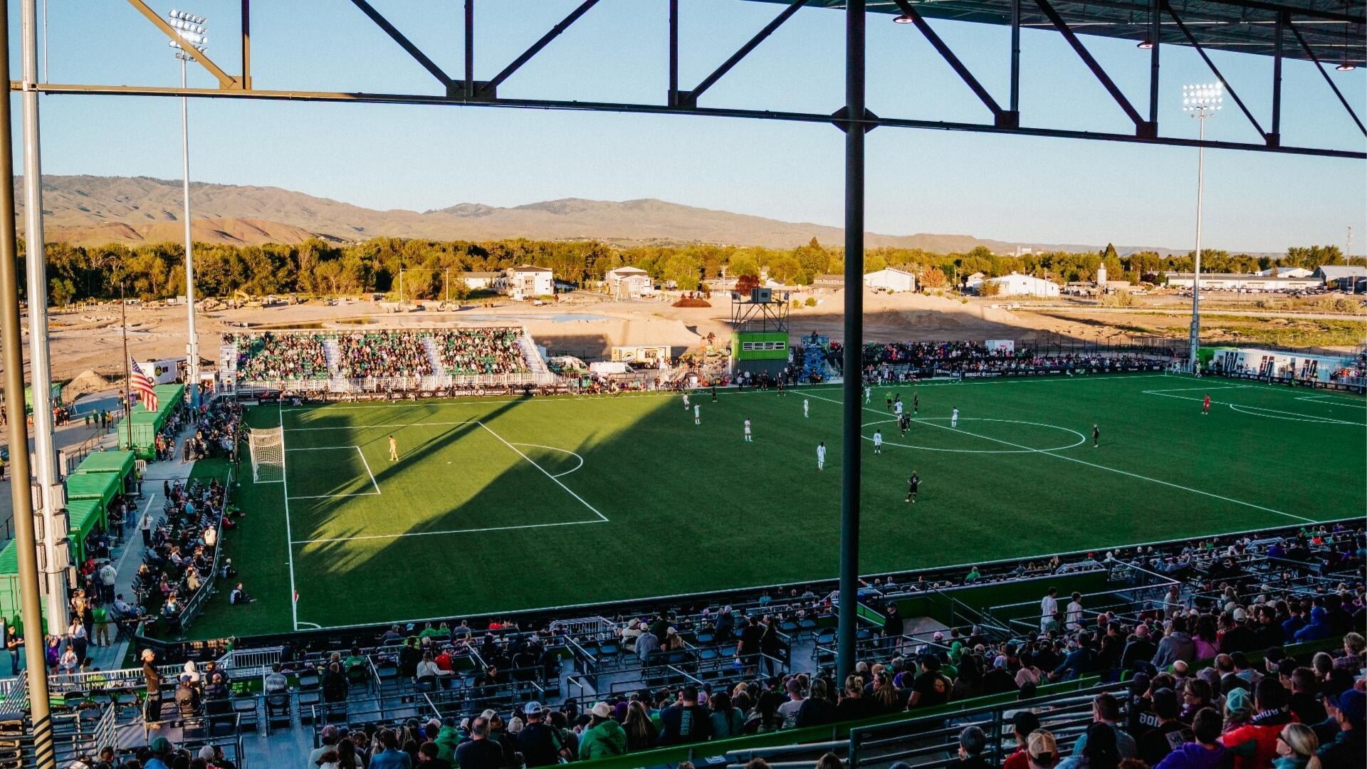 A soccer match is underway on a green turf field at an outdoor stadium, with players competing while spectators fill the stands against a backdrop of distant mountains.