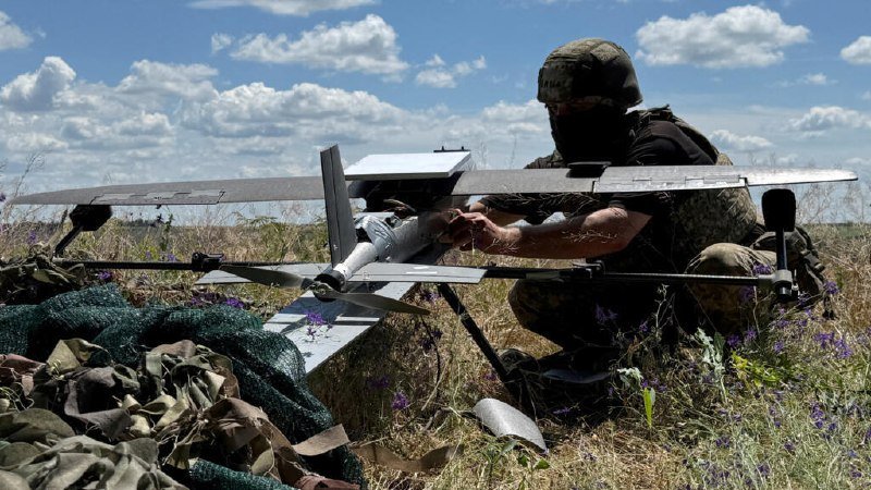 A masked person in camouflage gear prepares a small fixed-wing drone in a grassy field under a partly cloudy sky.