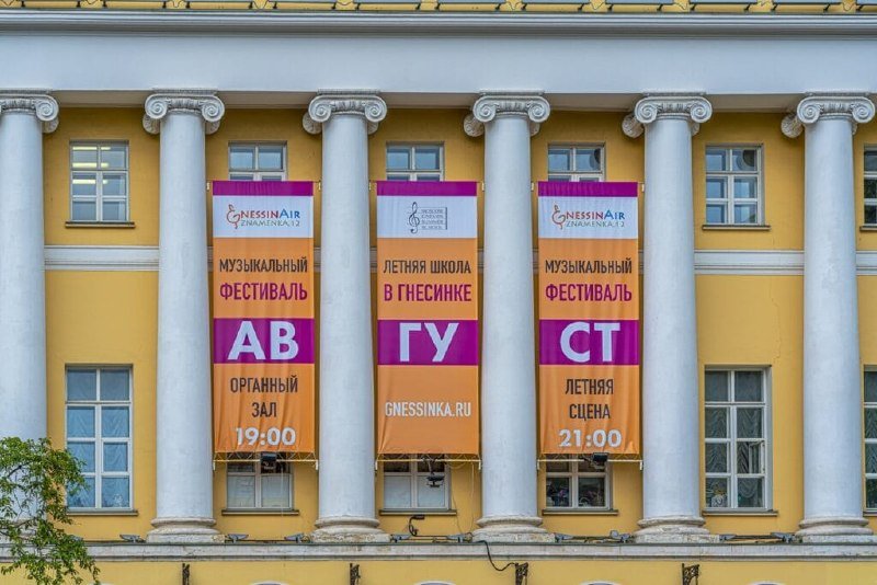 Three orange and magenta banners advertising the GnessinAir music festival hang between white columns on a yellow neoclassical building's facade.
