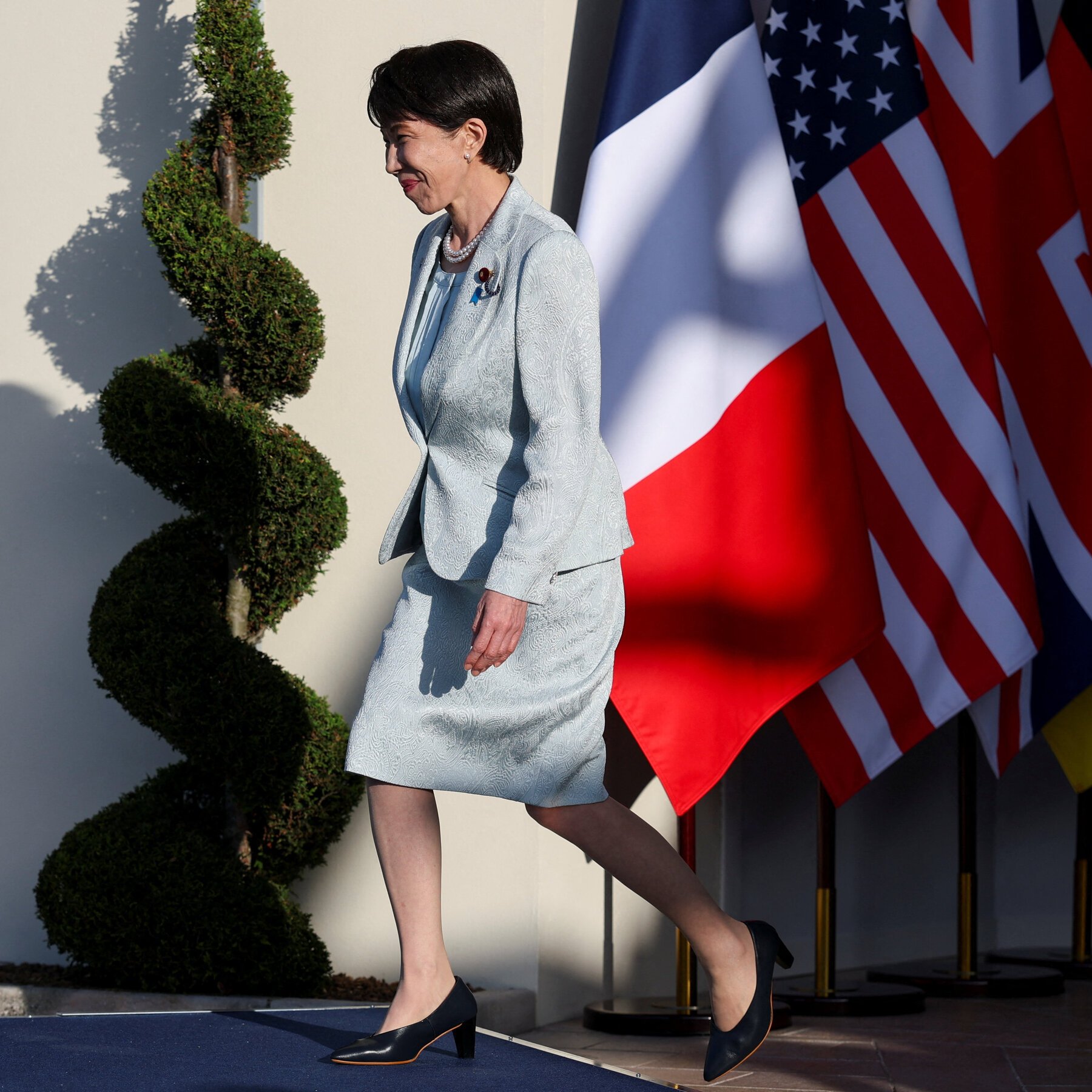 A woman in a light blue patterned skirt suit and pearl necklace walks past a spiral topiary and an American flag.