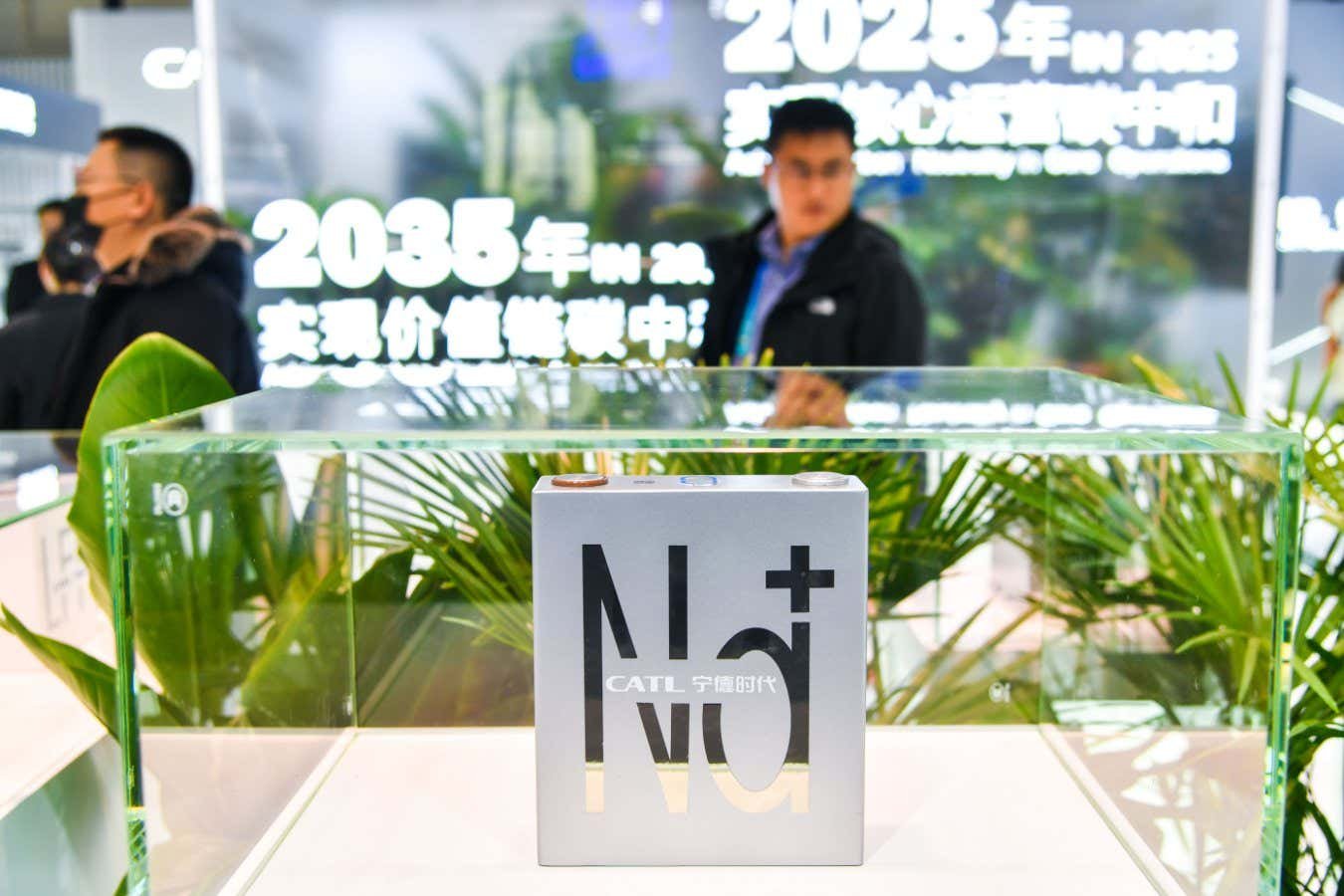 A Na+ battery cell branded "CATL" is displayed in a glass case, with blurred figures and Chinese signage referencing "2025" and "2035" visible behind it.