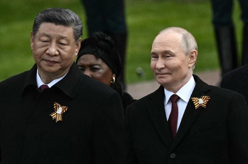 Two men in black suits and ties, each wearing an orange-and-black striped ribbon pin, stand side by side outdoors, smiling slightly.