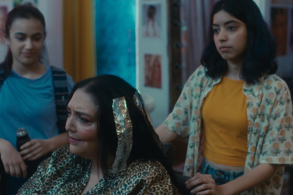 Three women appear together indoors, with one seated wearing a black headscarf and leopard-print outfit, flanked by two younger women, one in a blue top holding a bottle.