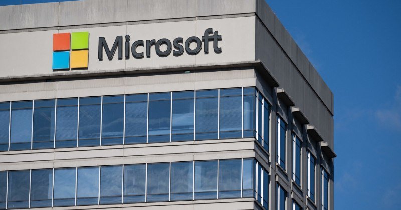 The Microsoft logo and name are mounted near the top of a modern multi-story building with rows of blue-tinted glass windows against a clear sky.