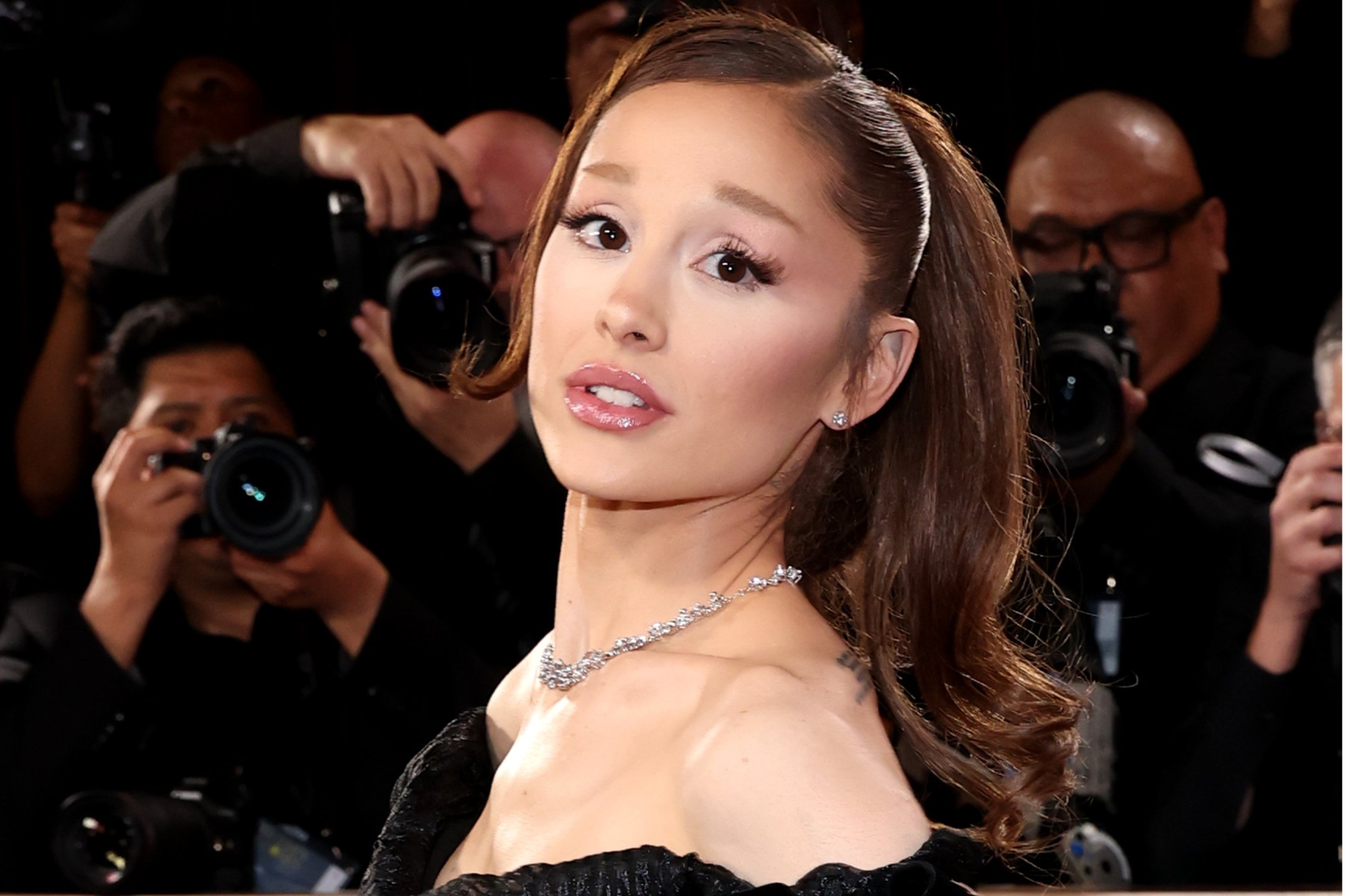 A woman in a black off-shoulder gown with a diamond necklace poses as photographers with large cameras take her picture against a dark backdrop.