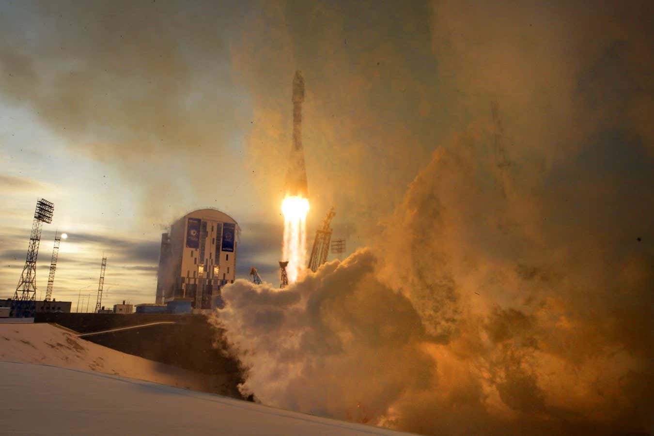 A rocket lifts off from a snowy launch pad, engulfed in billowing clouds of smoke and bright orange flame against a dusky sky.