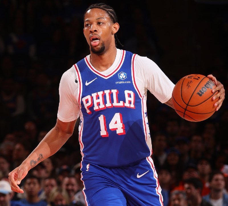 A basketball player wearing a blue "PHILA" jersey numbered 14 dribbles a basketball during a game against a blurred crowd backdrop.