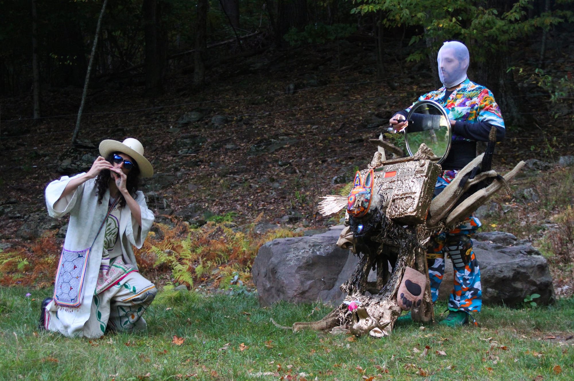 Two costumed performers pose outdoors on grass; one kneels in a white poncho, hat, and sunglasses, while the other wears a colorful patterned suit and a sheer white face covering, standing beside a gold-painted sculpture.