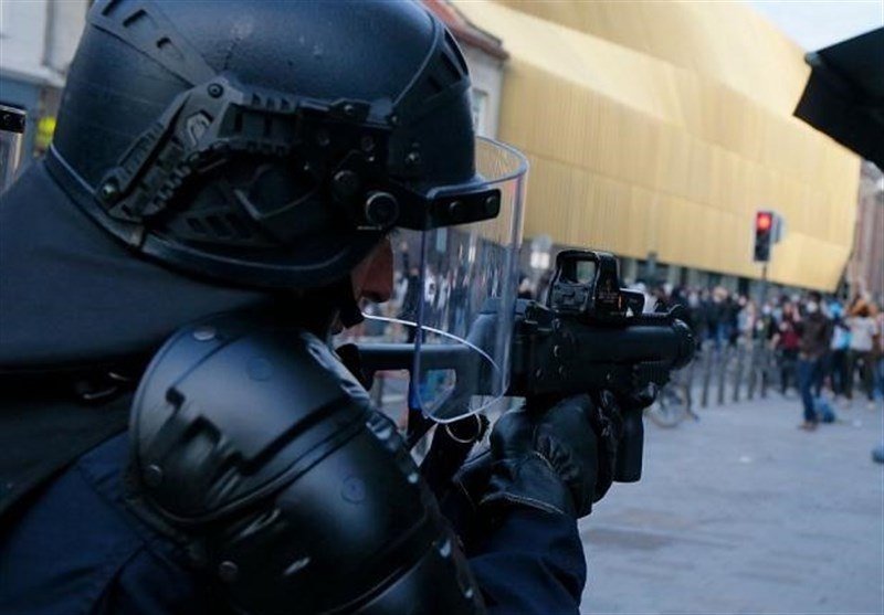 A person in riot gear aims a launcher-mounted weapon toward a crowd gathered in a city square near a traffic light.