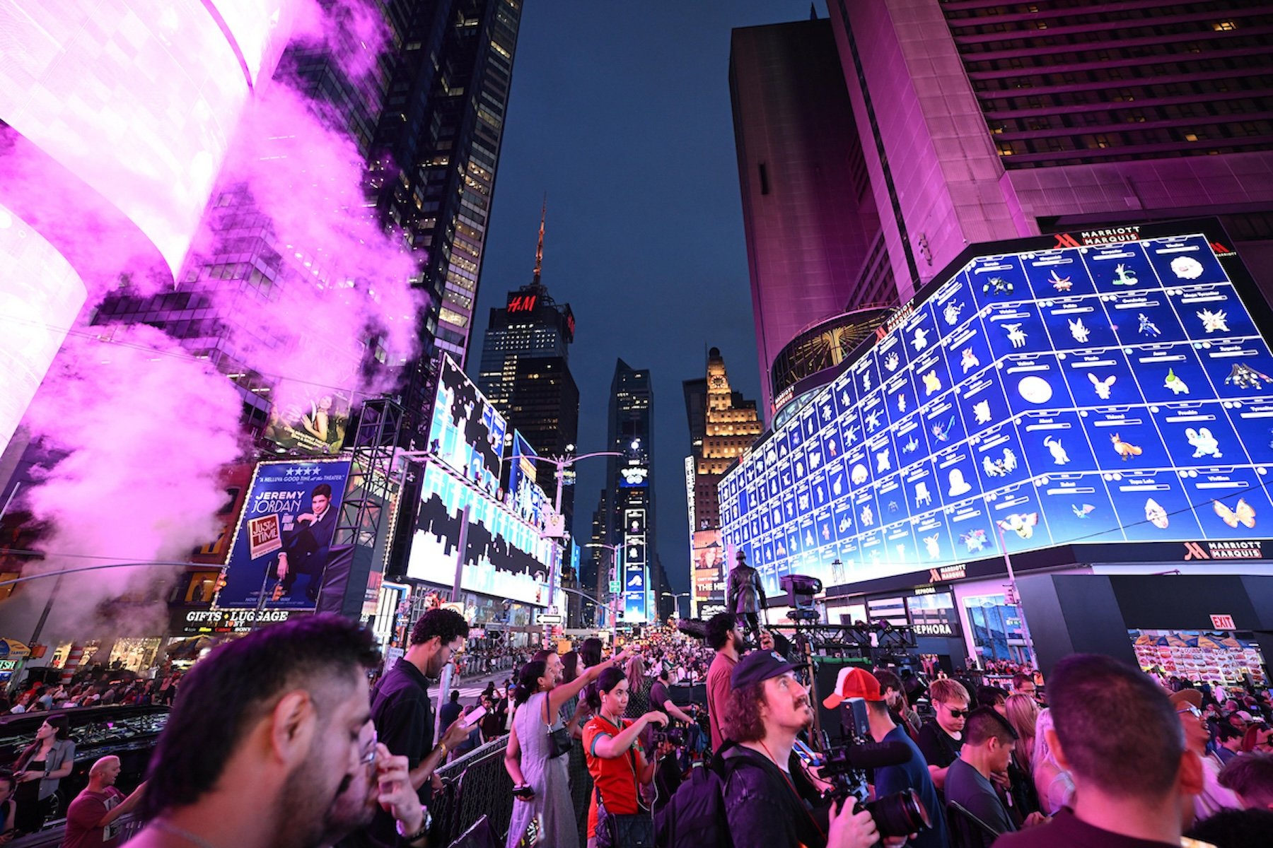 A crowd gathers in Times Square at dusk beneath illuminated billboards, as pink-lit steam rises from a vent on the left and photographers aim cameras toward the street.