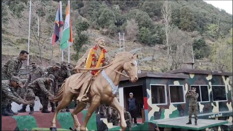 Uniformed personnel push a golden equestrian statue adorned with marigold garlands near flags and a camouflage-painted vehicle on a forested hillside.