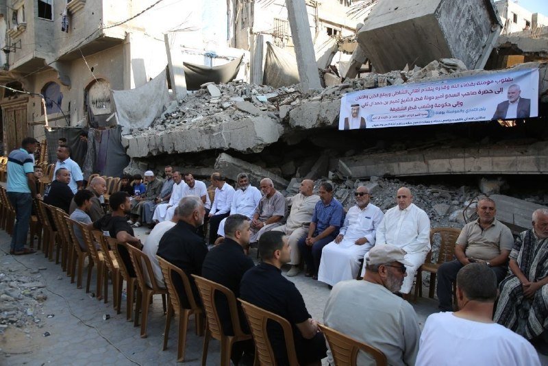 A group of men sit on plastic chairs gathered before a banner bearing Arabic text and two portraits, set against a backdrop of heavily damaged, collapsed concrete buildings.