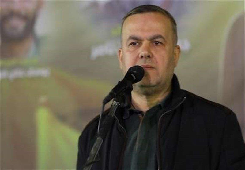 A man with short hair wearing a dark jacket stands at a microphone with a serious expression, a blurred graphic backdrop behind him.