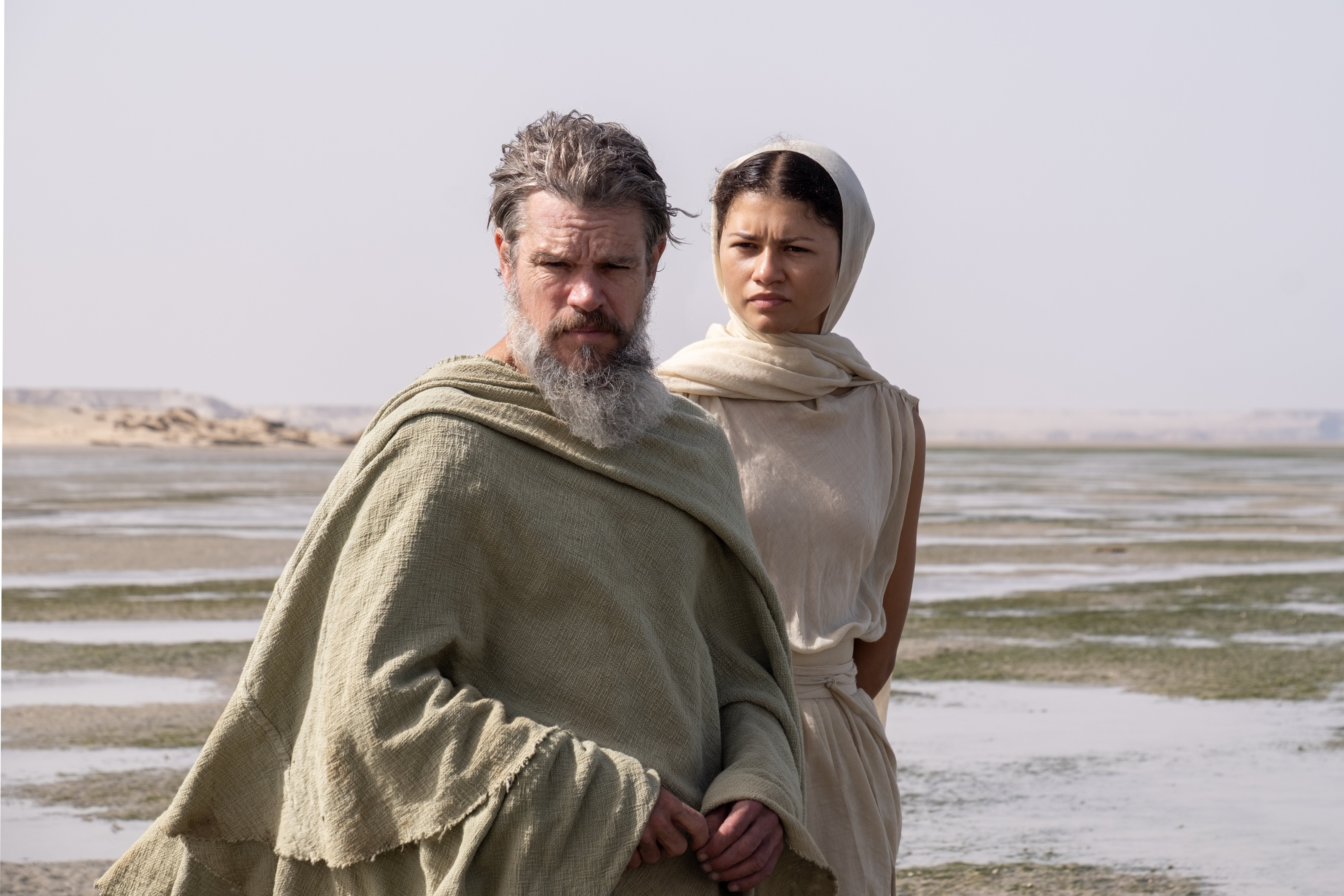 Two people in draped, earth-toned robes stand on a flat, wet shoreline under a hazy sky, gazing forward with serious expressions.