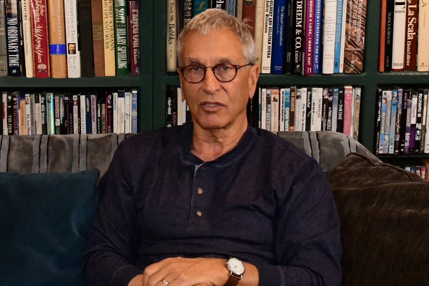 A gray-haired man wearing glasses and a dark button-up shirt sits on a couch in front of a bookshelf filled with various books and DVDs.