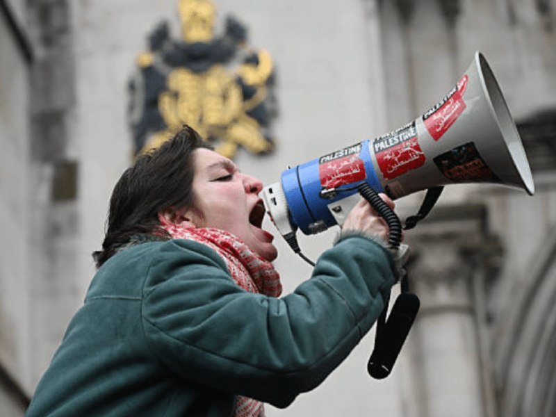 A woman with dark hair and a red-and-white scarf shouts into a megaphone labeled "PALESTINE," standing before a stone building displaying a large ornate crest.