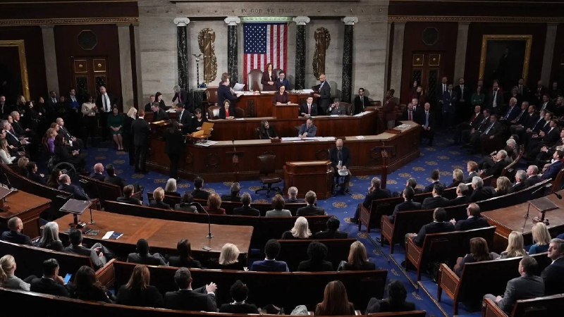 Lawmakers gather in a large legislative chamber with a U.S. flag and "IN GOD WE TRUST" inscribed above the speaker's rostrum, where officials preside over seated members.