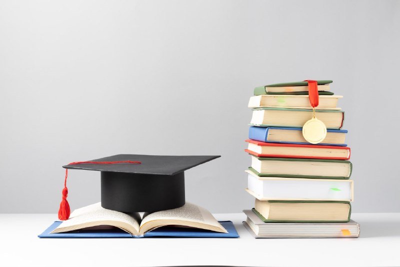 A black graduation cap rests on an open book beside a tall stack of colorful books with a gold medal against a plain white background.