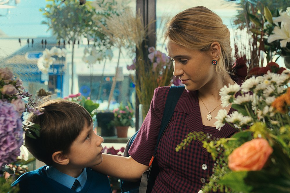 A blonde woman in a maroon checkered dress smiles down at a young boy in a blue sweater among colorful flower bouquets inside a shop.