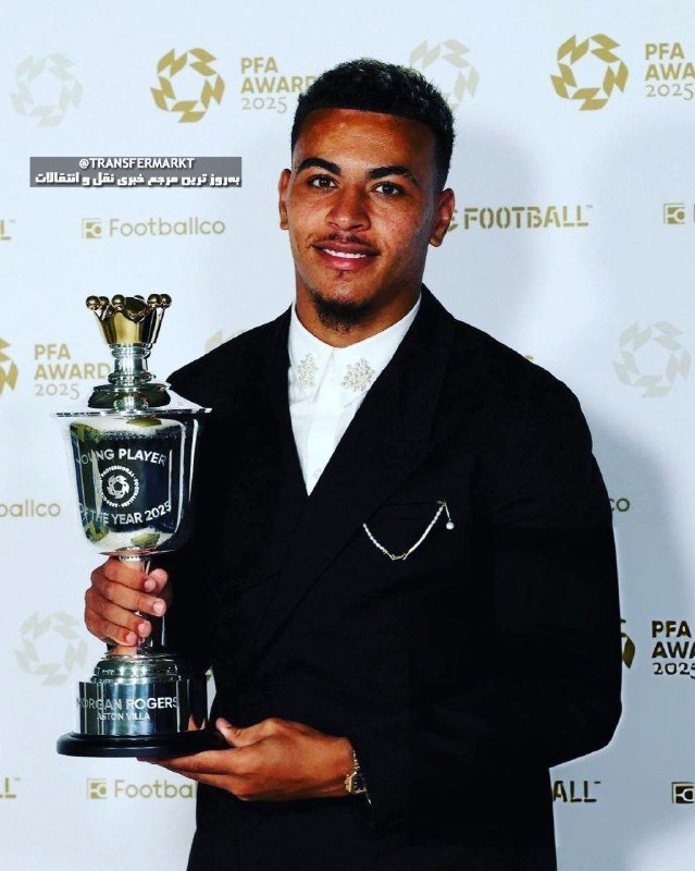 A young man in a black suit with a chain brooch smiles while holding a silver trophy labeled "Young Player of the Year 2025" at a PFA Awards backdrop.