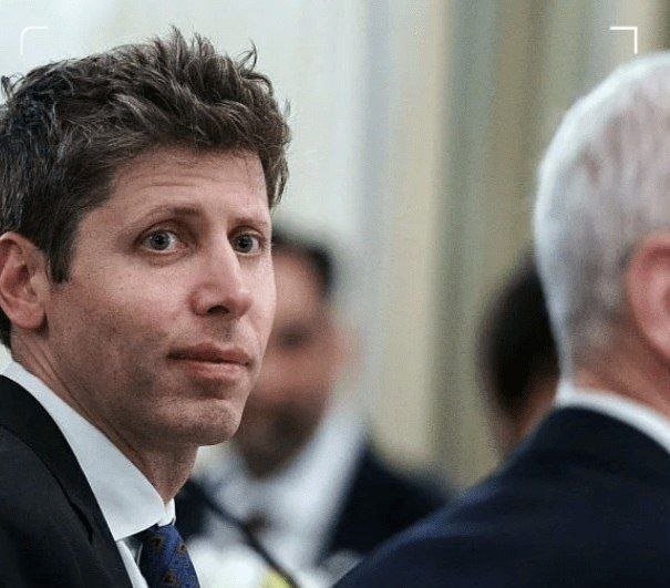 A dark-haired man in a dark suit, white shirt, and patterned tie looks to the side, with another gray-haired man partially visible in the foreground.