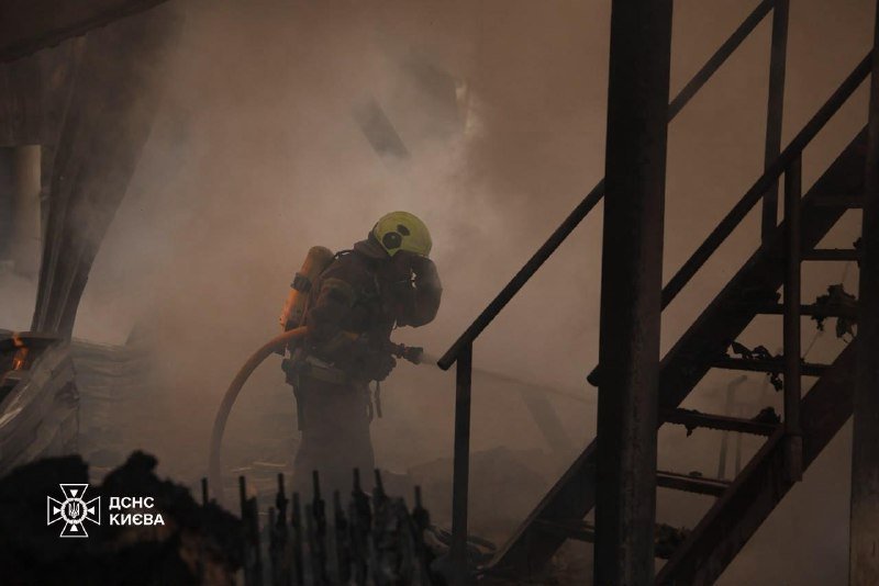 A firefighter in full gear and helmet works amid heavy smoke beside a metal staircase in a fire-damaged structure, with a "ДСНС КИЄВА" logo visible.