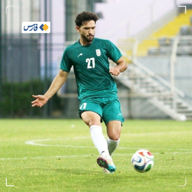 A bearded soccer player wearing a green jersey with the number 27 dribbles a ball on a grass field.