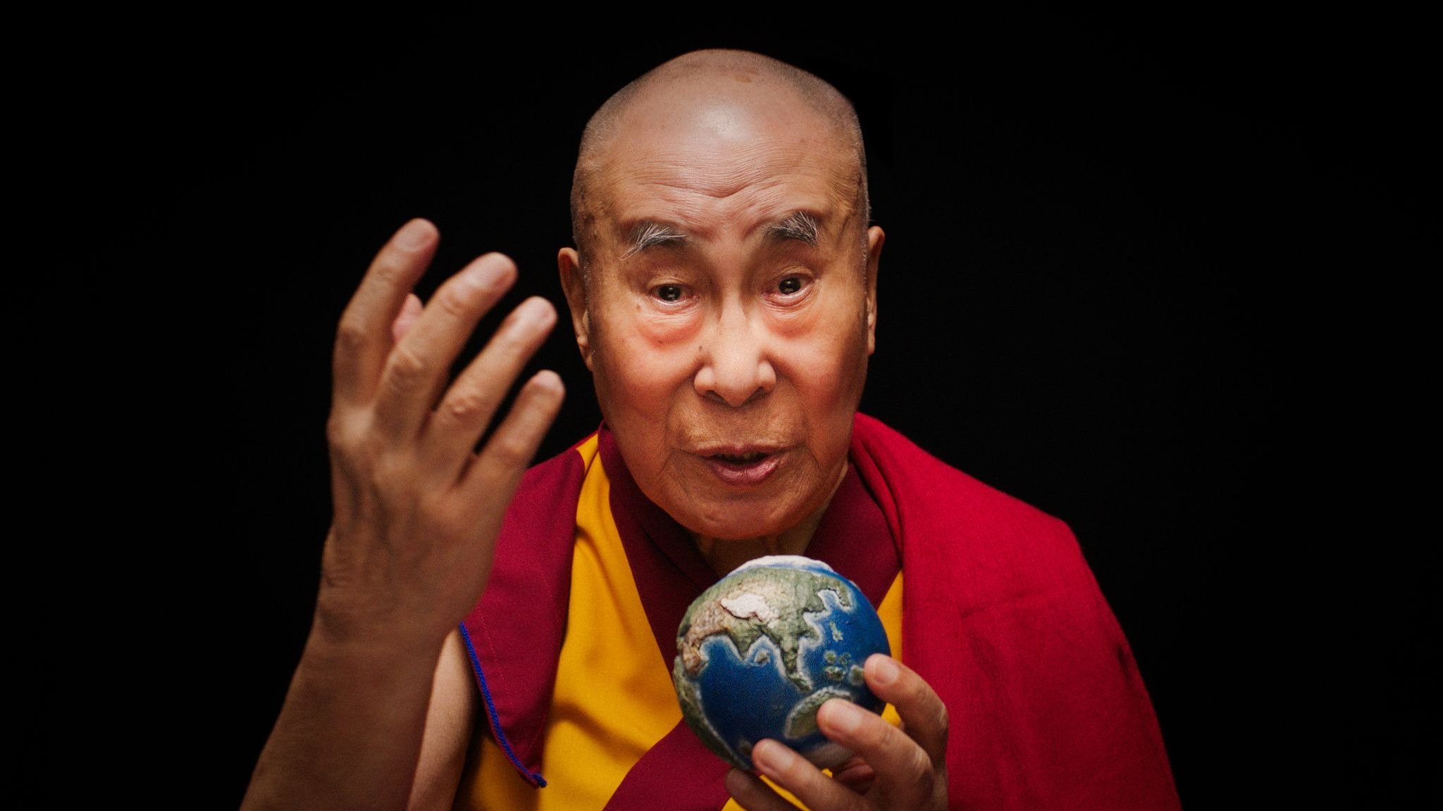 A bald elderly man in red and yellow robes holds a small globe in his hands and gestures with one raised hand against a black background.