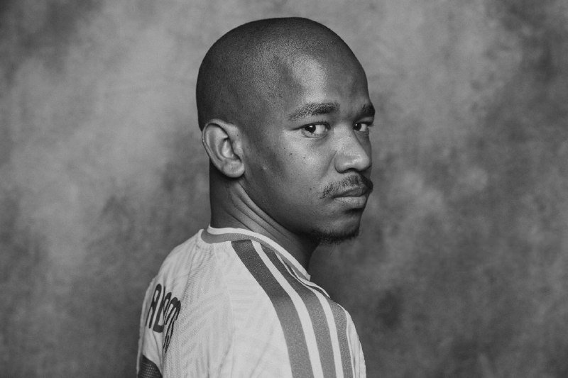 Black-and-white portrait of a man with a short mustache wearing a striped athletic jersey, looking over his shoulder against a mottled gray backdrop.