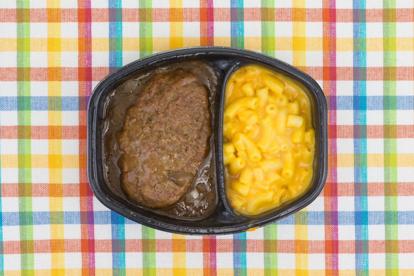 A black plastic divided tray containing a meat patty in brown gravy and macaroni and cheese sits on a multicolored plaid tablecloth.