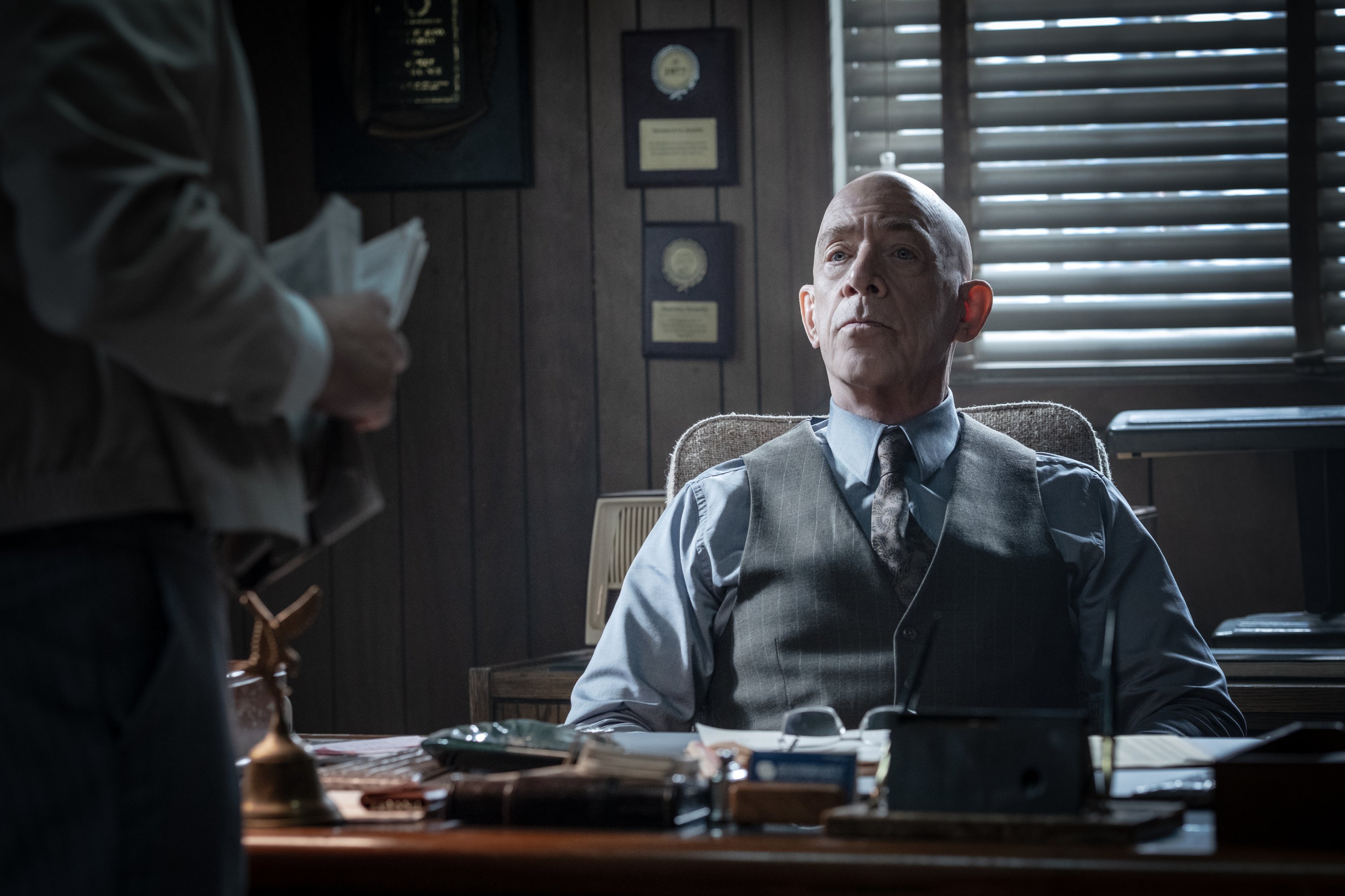 A bald man in a vest and tie sits at a cluttered office desk, looking up toward a standing person holding papers, with wall plaques and shuttered windows behind him.