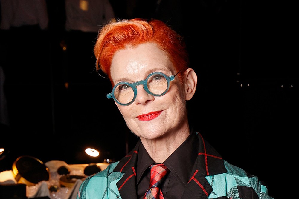 A woman with short bright orange hair, teal round glasses, red lipstick, and a plaid blazer looks toward the camera against a dark background.