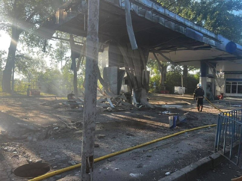 Firefighter in uniform hoses debris beneath a severely damaged, partially collapsed commercial building's metal canopy at sunset.