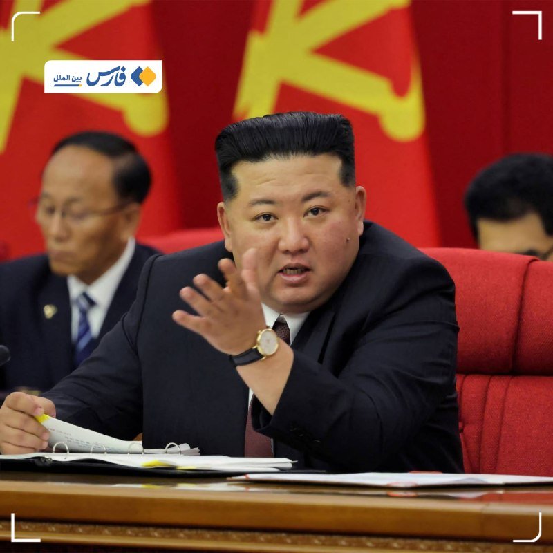 A man in a dark suit gestures with his right hand while seated at a table with documents, with other suited individuals and a red flag with yellow symbols behind him.