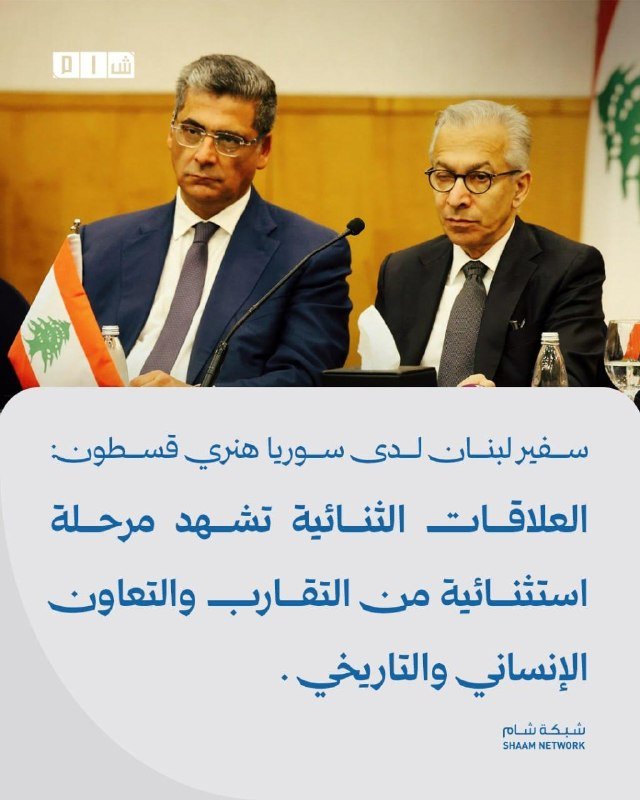 Two men in suits and glasses sit at a table with microphones, beside a Lebanese flag, beneath Arabic text and the Shaam Network logo.