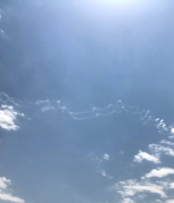 Wavy contrails stretch across a bright blue sky dotted with scattered white clouds.