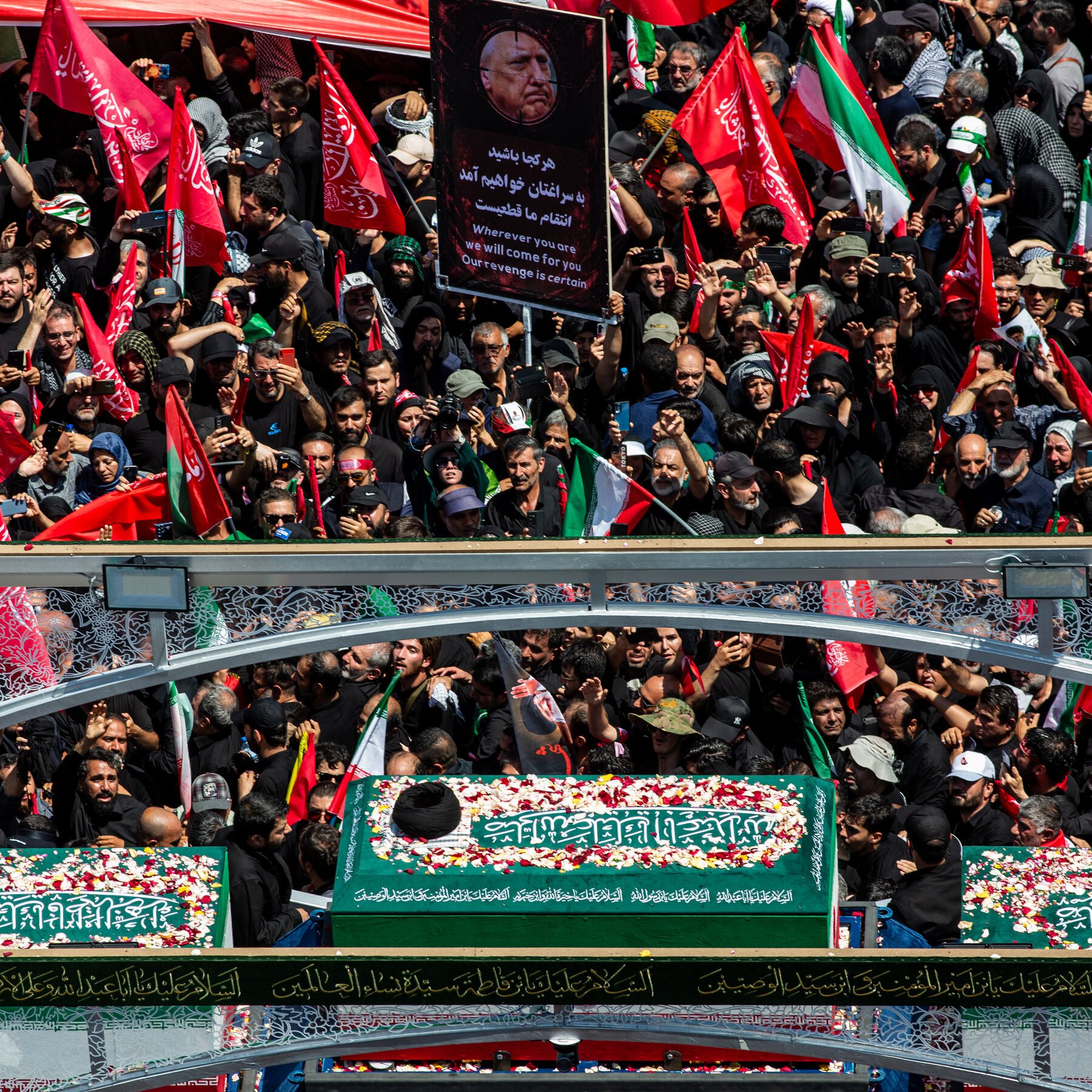 A large crowd, many dressed in black, gathers around green flower-adorned coffins bearing Arabic and Persian inscriptions, waving red and Iranian flags while holding a portrait placard reading "Wherever you are we will come for you, Our revenge is certain."