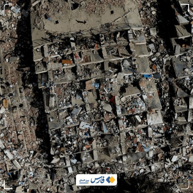 An aerial view shows a densely built area of collapsed buildings and rubble-strewn streets, with an Arabic news logo and watermark overlaid at the bottom.