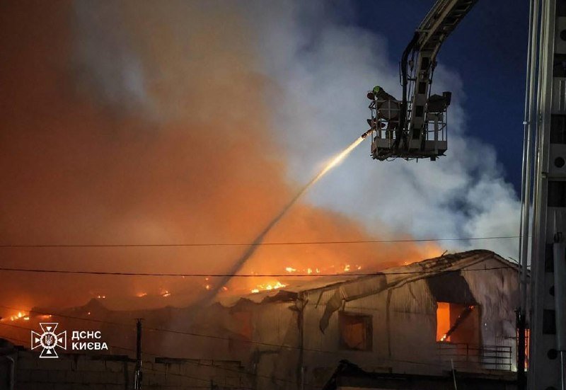 Firefighters in an elevated ladder bucket spray water onto a large burning building engulfed in flames and heavy smoke at night, with "ДСНС КИЄВА" watermarked at bottom left.