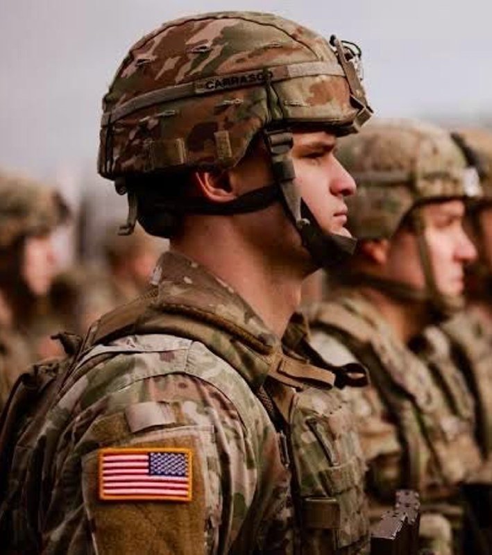 Soldiers in camouflage uniforms and helmets stand in formation, with a U.S. flag patch visible on the foreground soldier's sleeve.