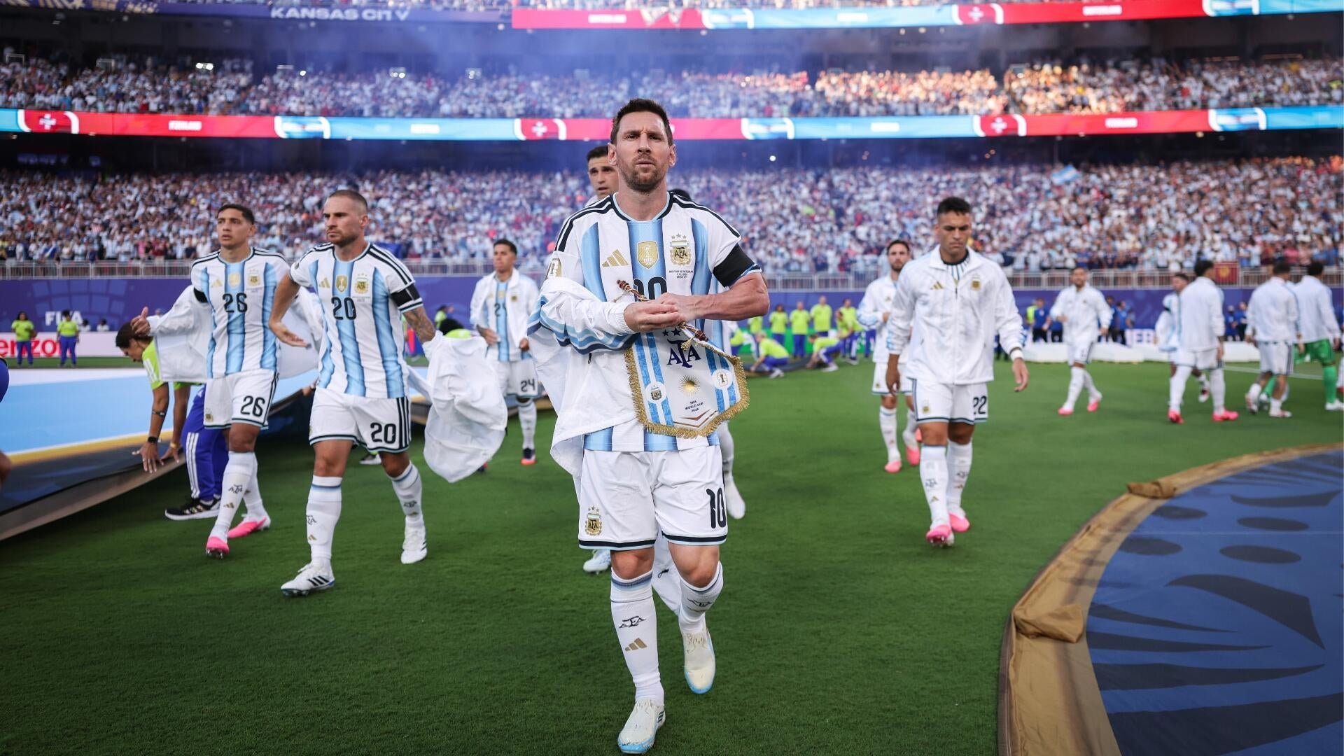 Soccer players in light blue and white striped jerseys walk onto the field carrying a pennant, with "KANSAS CITY" visible on the stadium signage above a packed crowd.