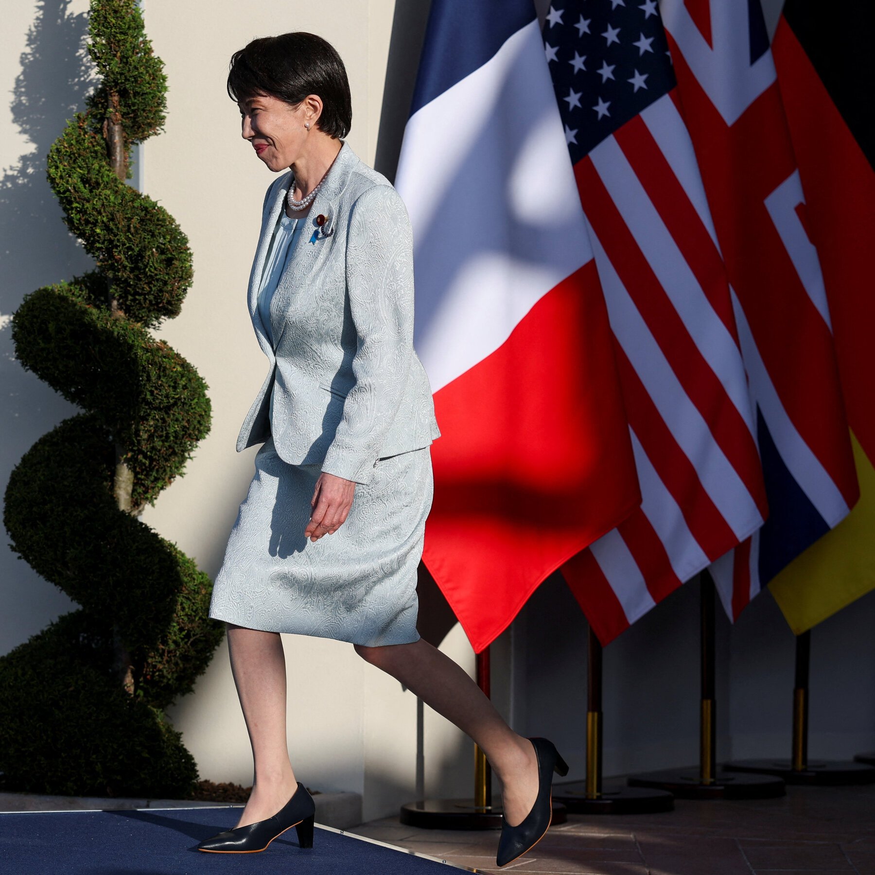 A woman in a light blue skirt suit walks past spiraled topiary and U.S. and Japanese flags.