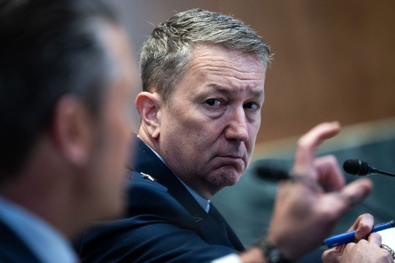 A man in a dark uniform sits at a hearing with a microphone visible, gesturing with his hand while looking sideways toward another person partially in frame.