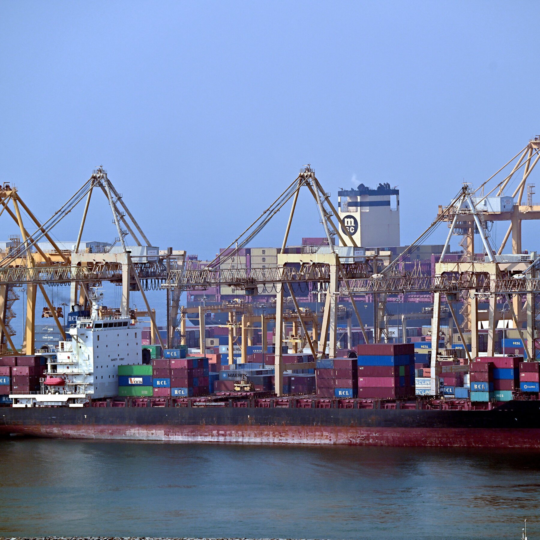 A large container ship loaded with multicolored shipping containers is docked at a port, with massive cranes and stacked containers visible in the background.