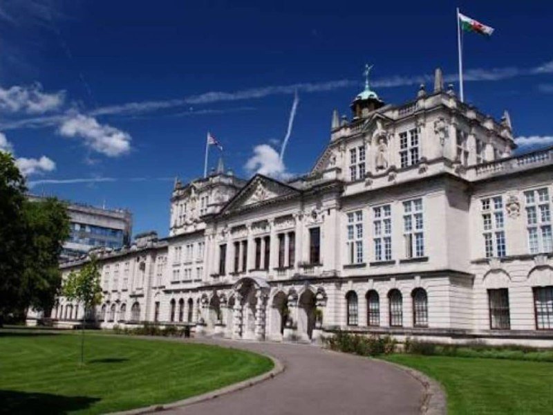 A grand, ornate stone building with arched windows and a central tower flies a flag under a blue sky with scattered clouds, surrounded by green lawn and a curved pathway.