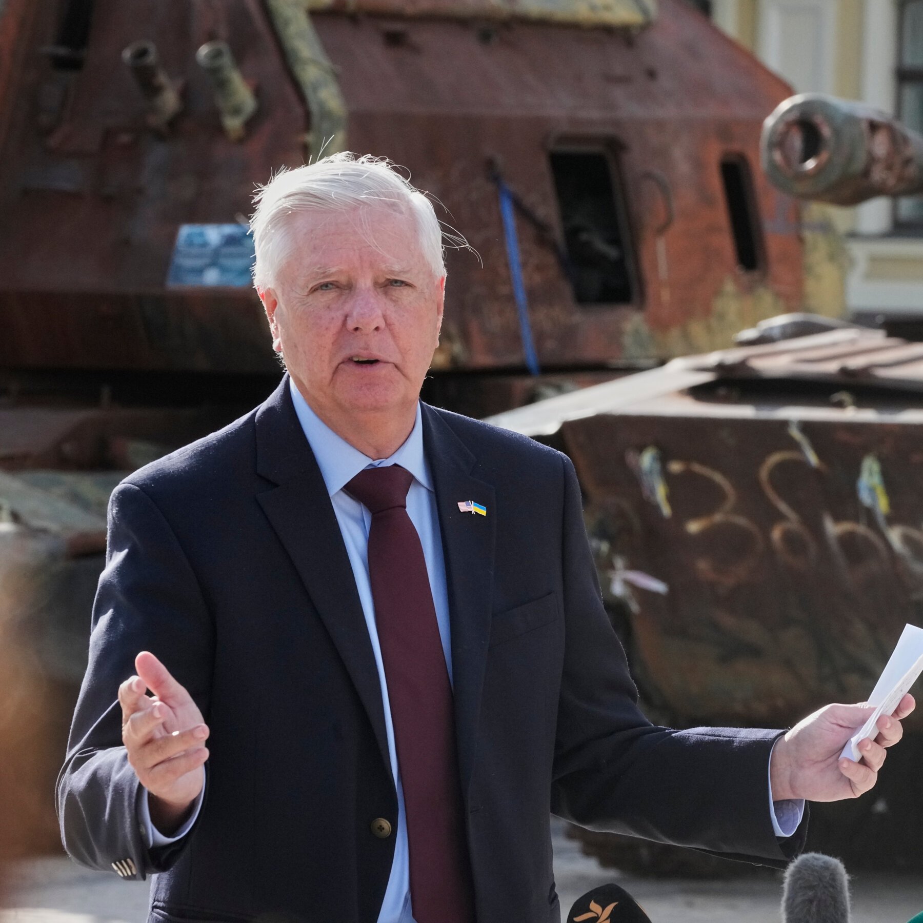 A white-haired man in a dark suit with a U.S. and Ukrainian flag lapel pin speaks and gestures in front of a rusted tank.