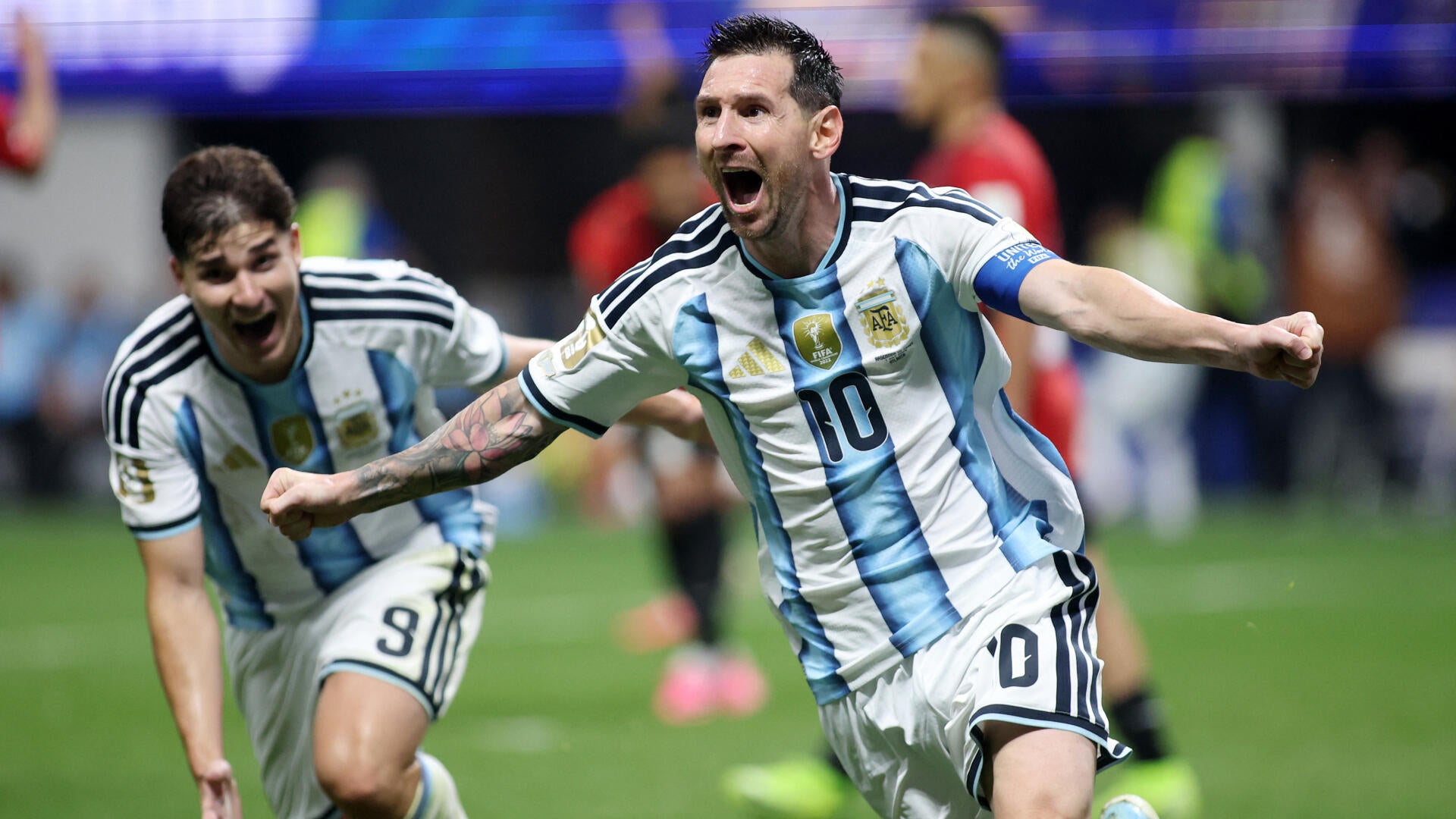 A soccer player in a black and blue jersey with the number 10 celebrates with arms outstretched on the field before a stadium crowd.