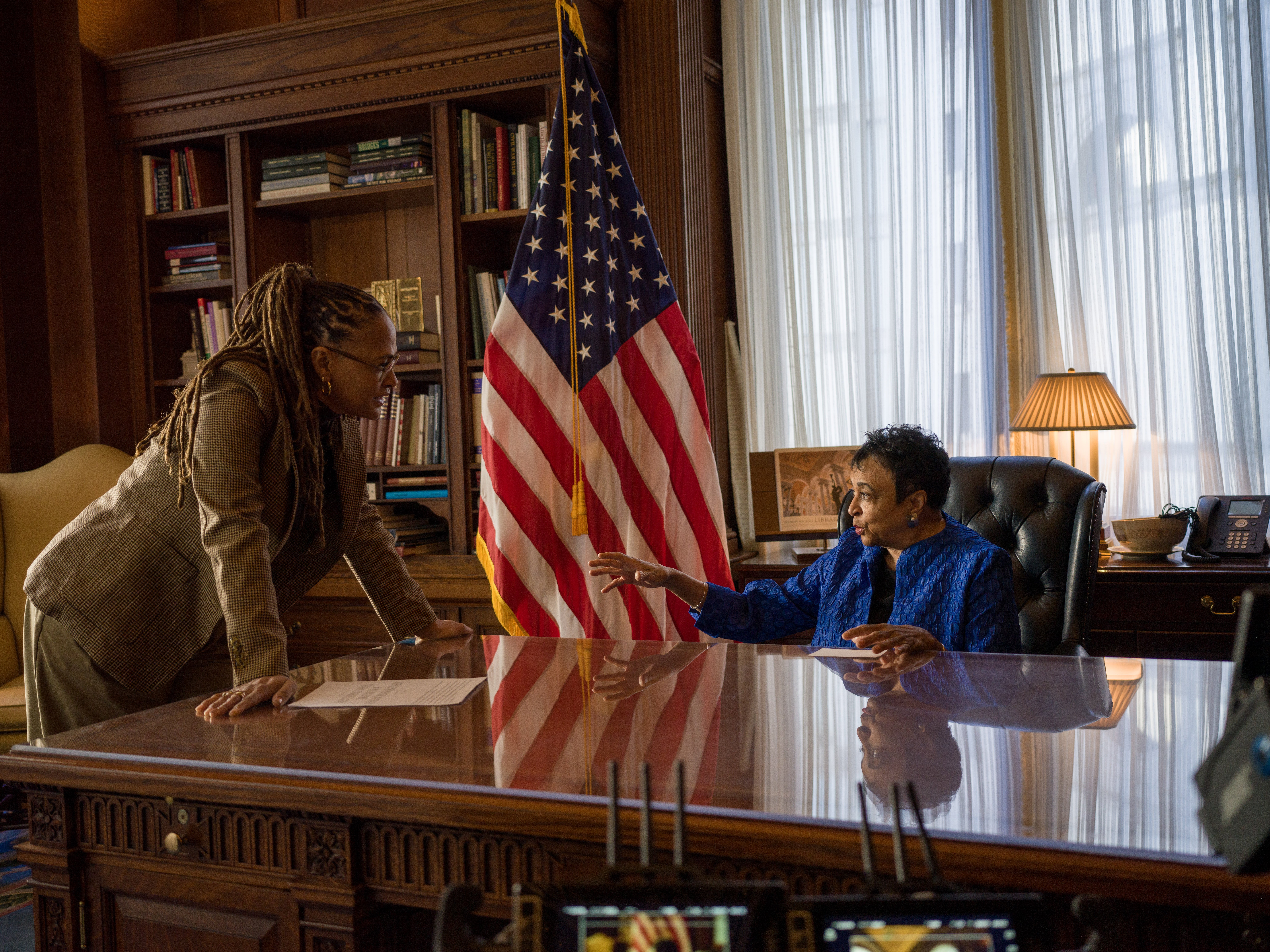 Two women converse across a polished wooden desk in an office, with a U.S. flag, bookshelves, and sheer curtains visible in the background.