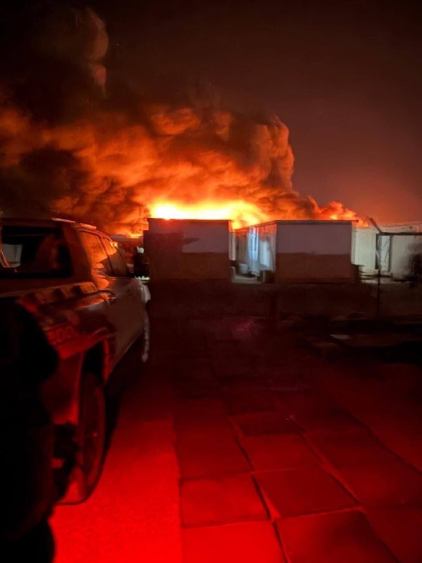 Large flames and thick smoke engulf warehouse structures at night, with parked vehicles and tiled flooring visible in the foreground.