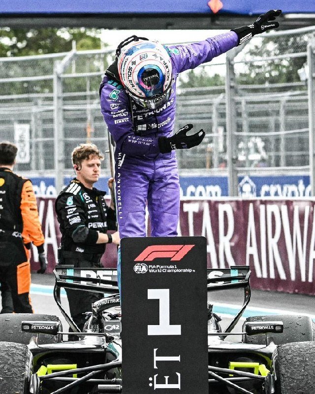 A driver in a purple racing suit stands atop a Mercedes Formula 1 car, waving from a "#1" podium marker after a race.